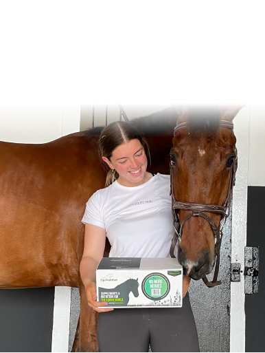 A smiling woman in a stable holds a box of supplements next to her brown horse.