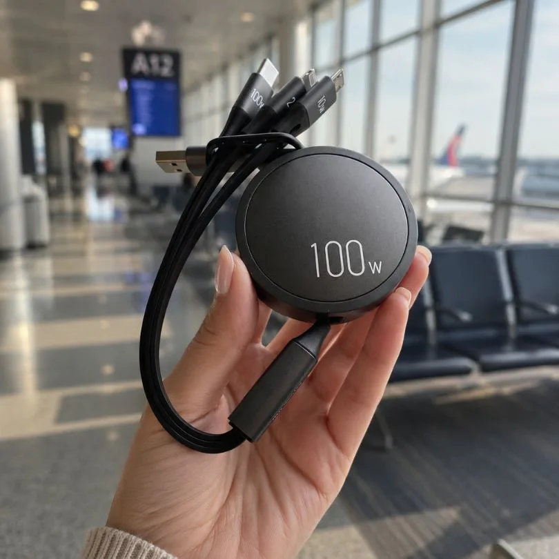 A hand holding a black, round, 100W multi-port charging cable in an airport terminal.