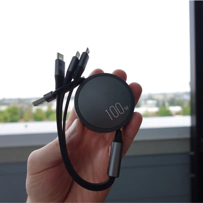 A hand holds a black, circular 100w multi-connector charging cable against a blurred outdoor background.