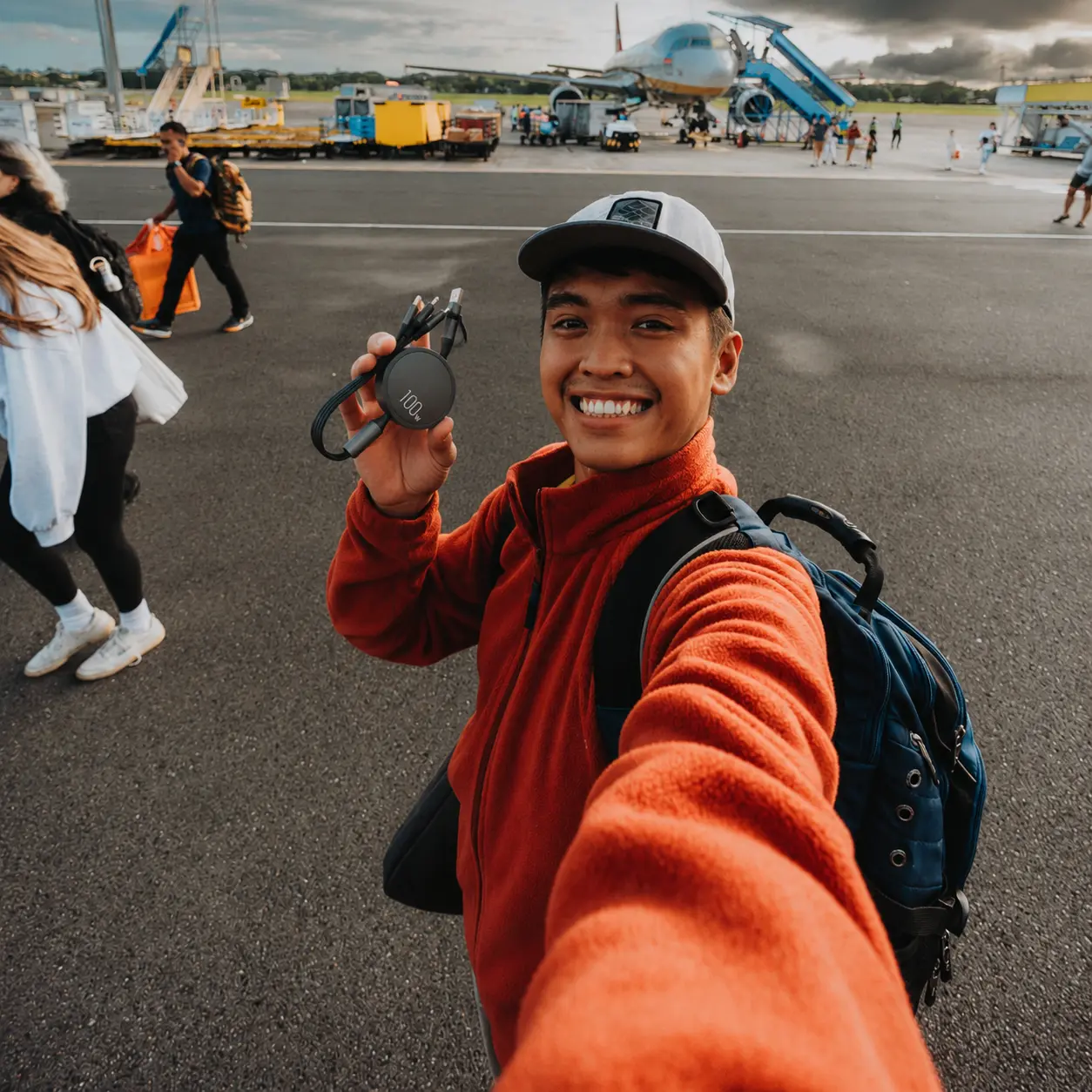 A smiling man takes a selfie on an airport tarmac, holding a charger, with an airplane behind him.
