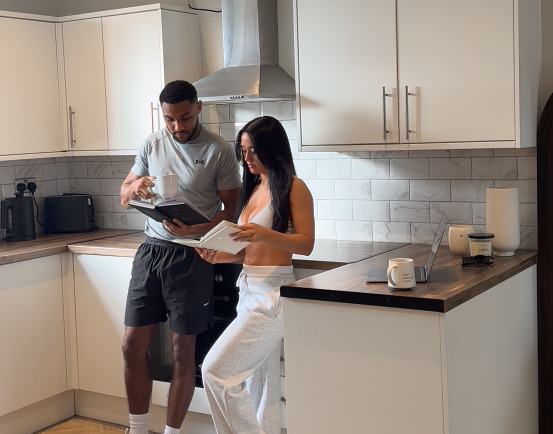 A man and a woman stand in a kitchen, looking at books, while the man holds a mug.