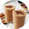 Two glasses of chocolate smoothie topped with chocolate shavings, on a table with wooden spoons and dates.