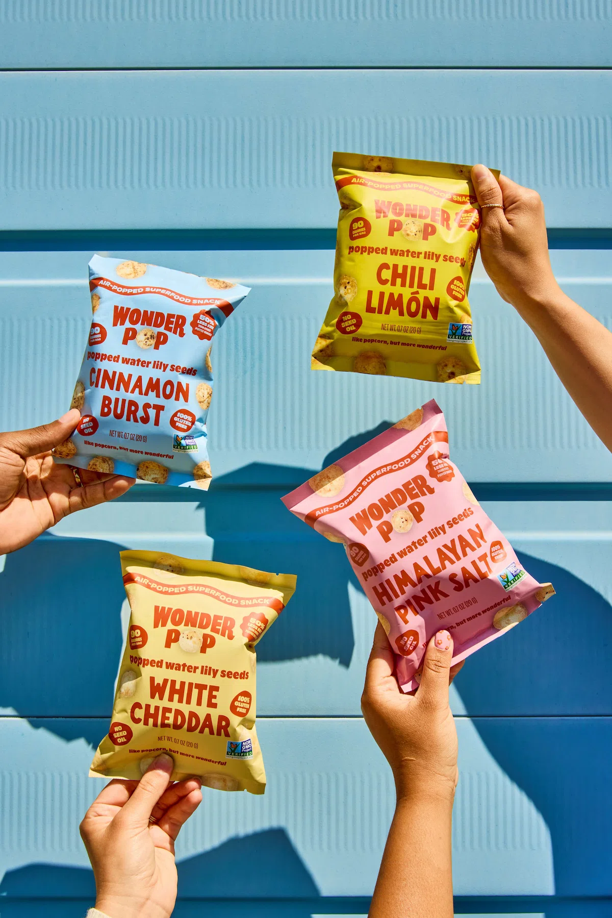 Four hands hold up different flavors of Wonder Pop popped water lily seed snacks against a blue background.