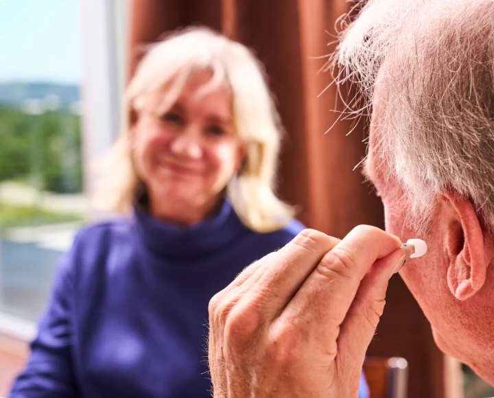 An older man inserts a hearing aid into his ear as a woman watches him and smiles.