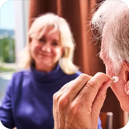 Person adjusting a hearing aid while another smiles in the background.