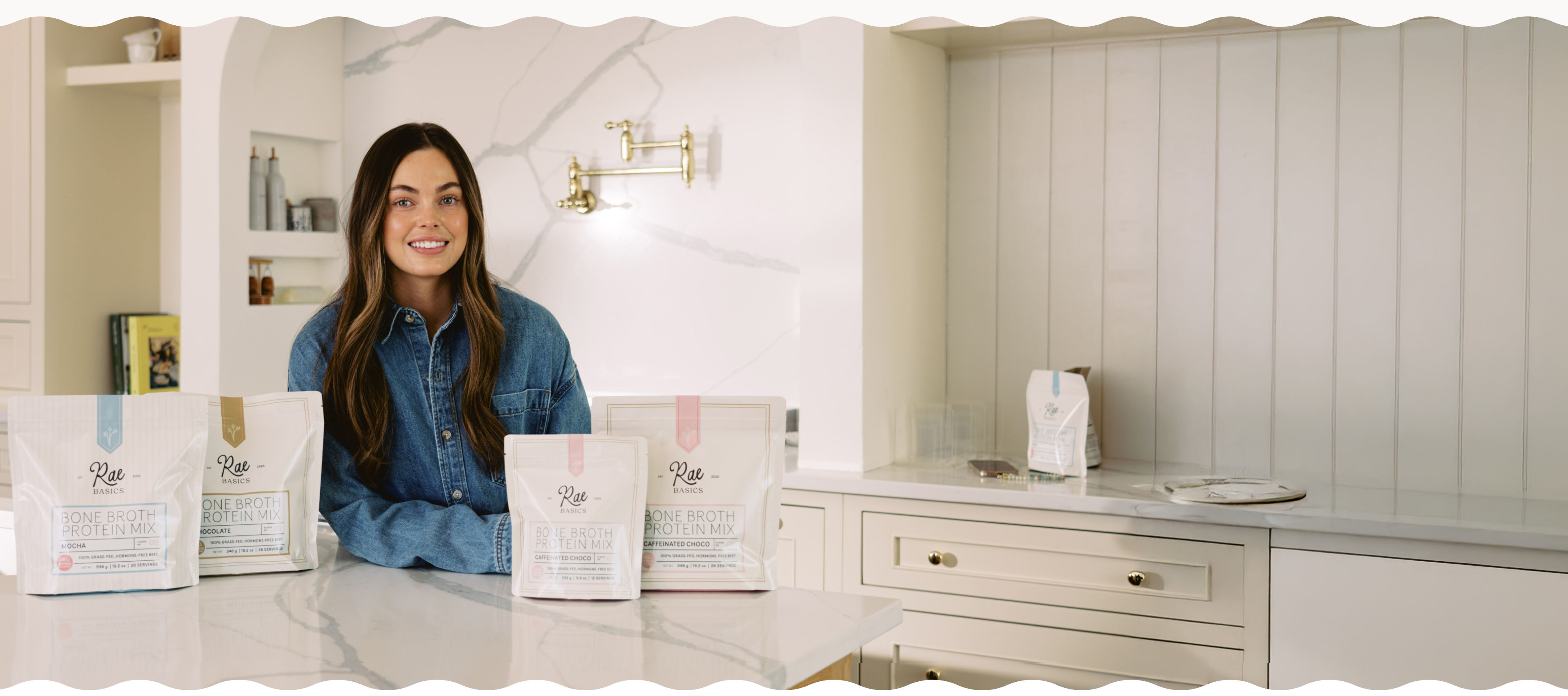 A woman in a denim shirt smiles behind pouches of Rae Basics protein mix on a kitchen counter.