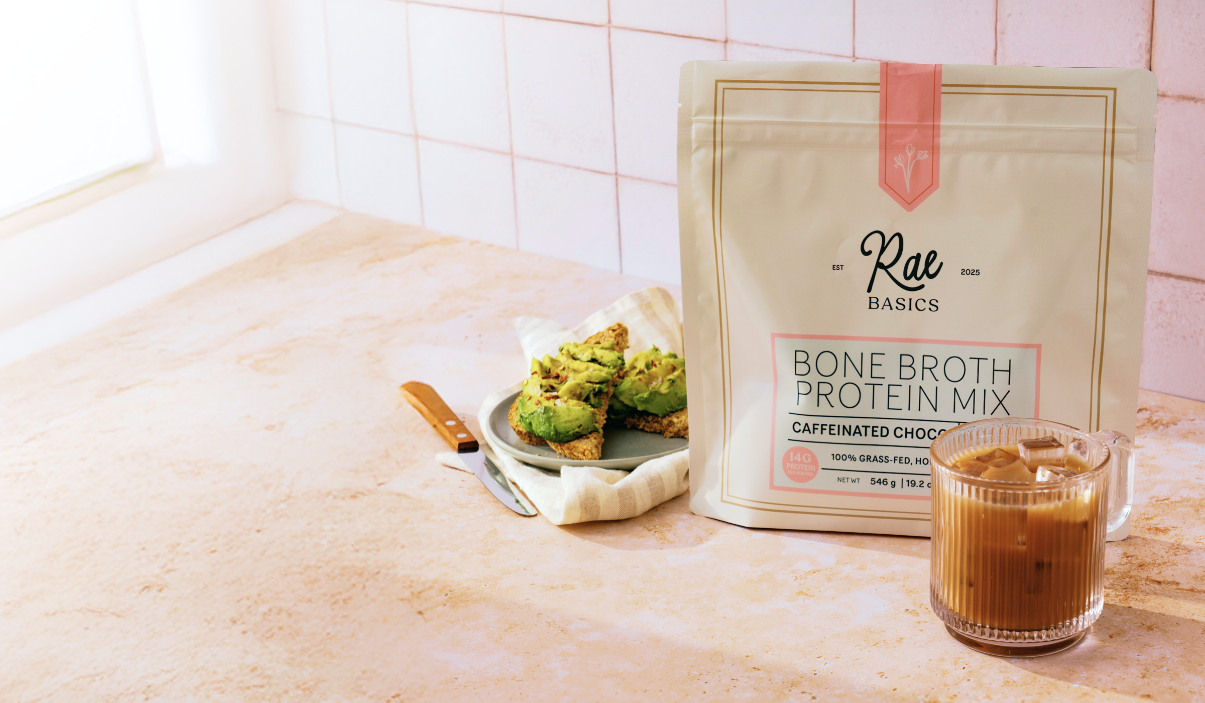 A bag of Rae Basics Bone Broth Protein Mix on a kitchen counter with avocado toast and iced coffee.