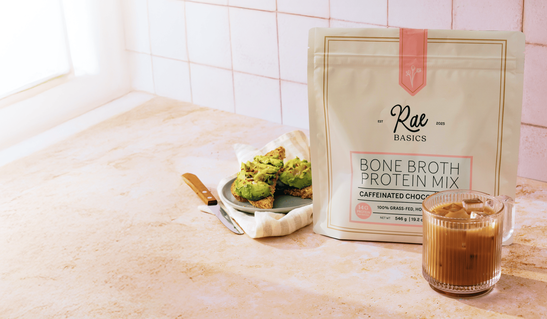 A bag of Rae Basics Bone Broth Hot Chocolate on a kitchen counter with avocado toast and iced coffee