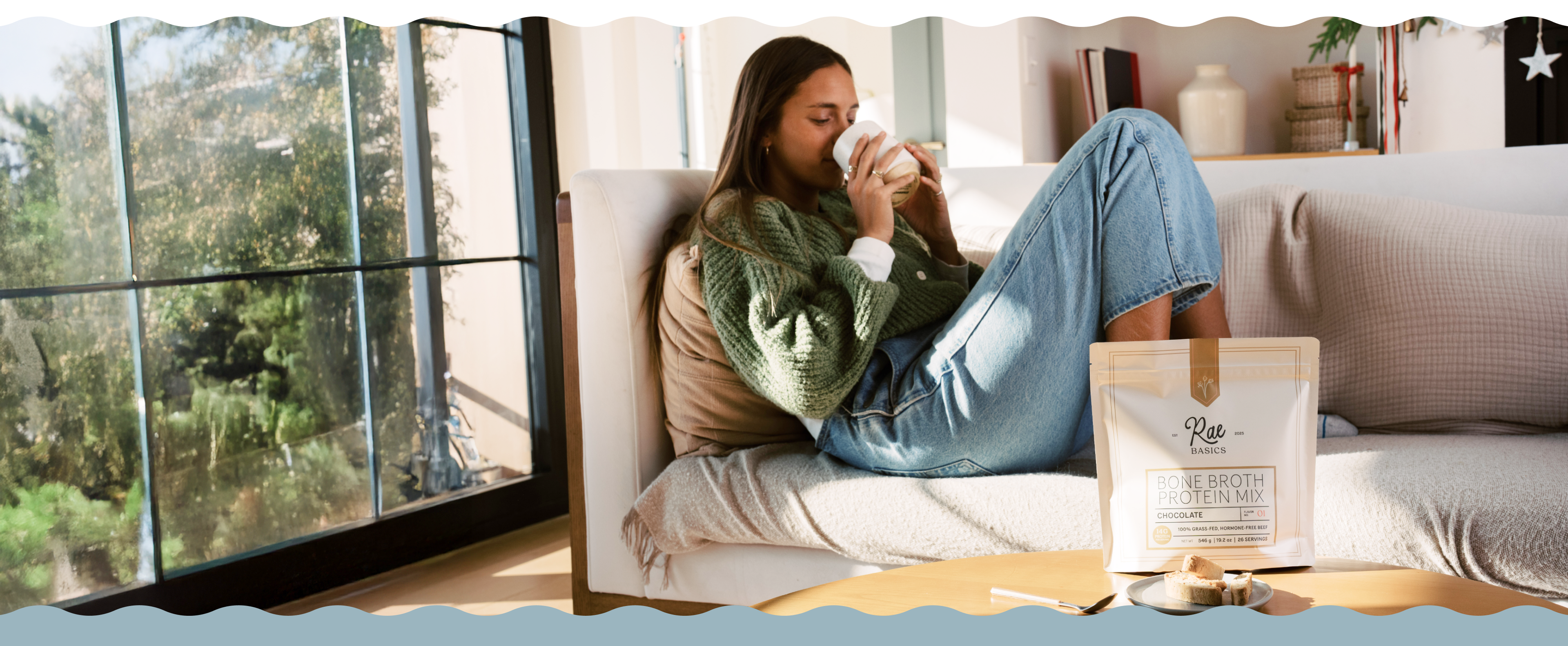 A woman in a green sweater and jeans sits on a couch, drinking from a mug.