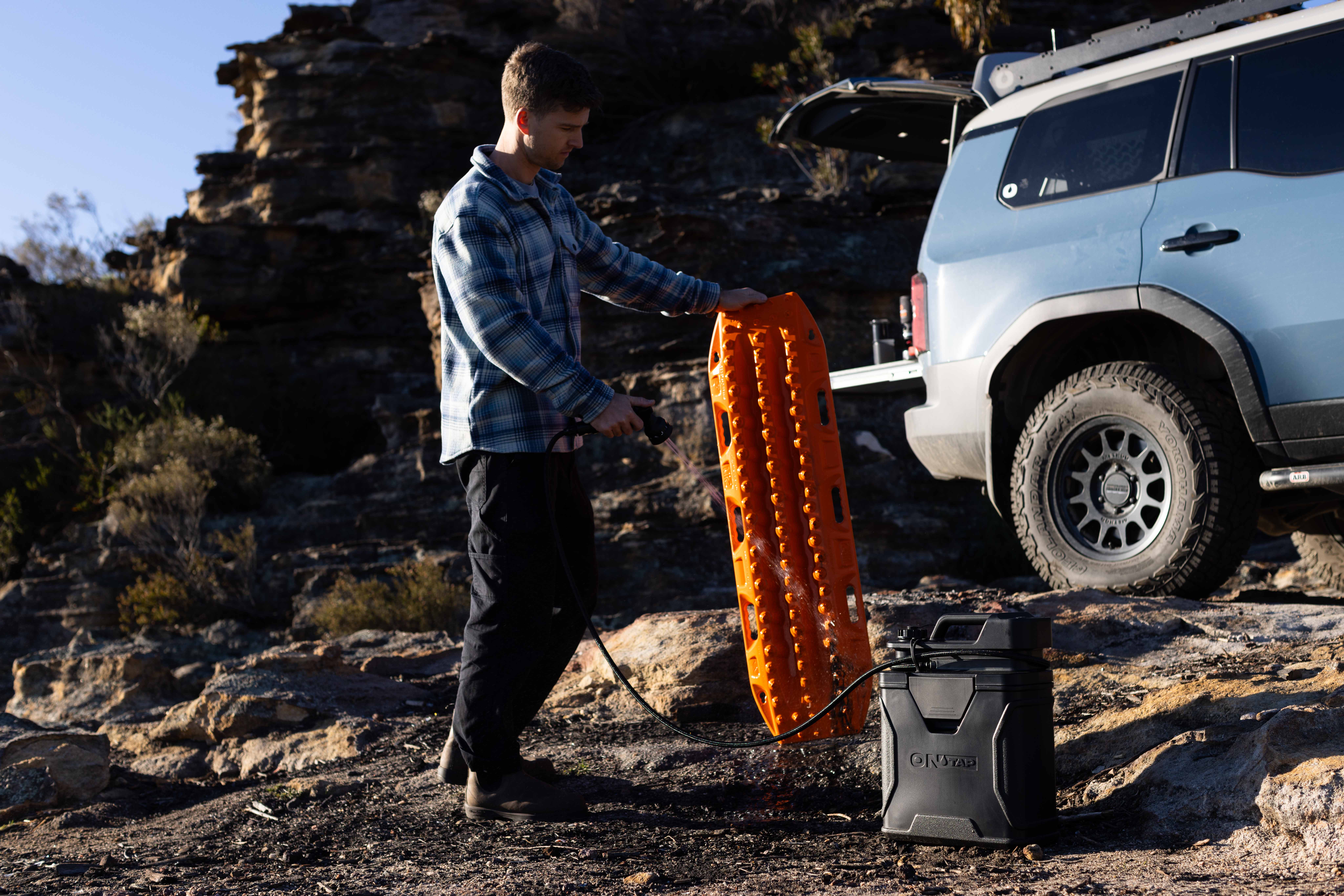 A man sprays water on an orange traction board next to a blue SUV in a rocky area.