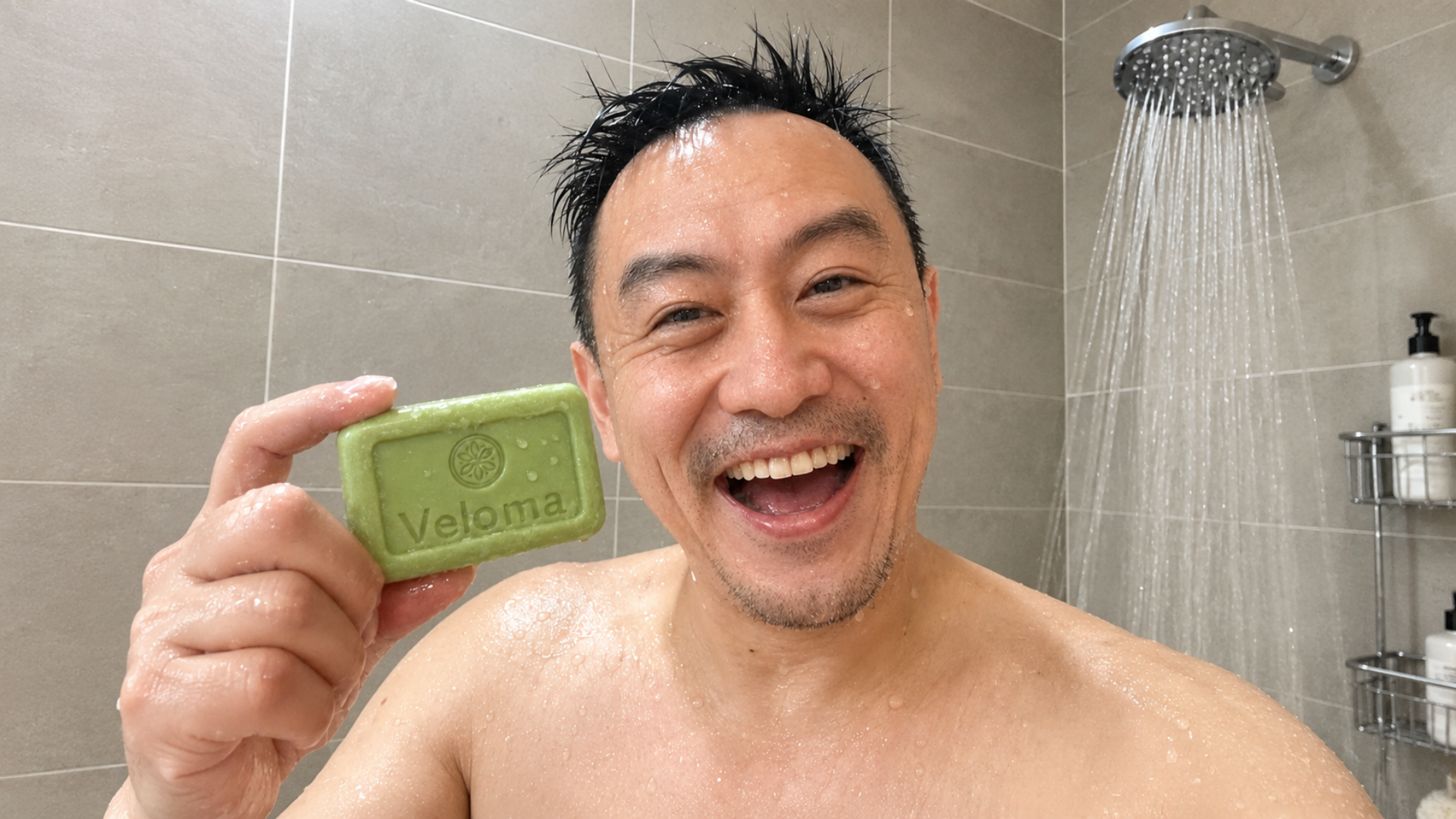 A smiling man with wet hair holds up a green bar of soap in a tiled shower.