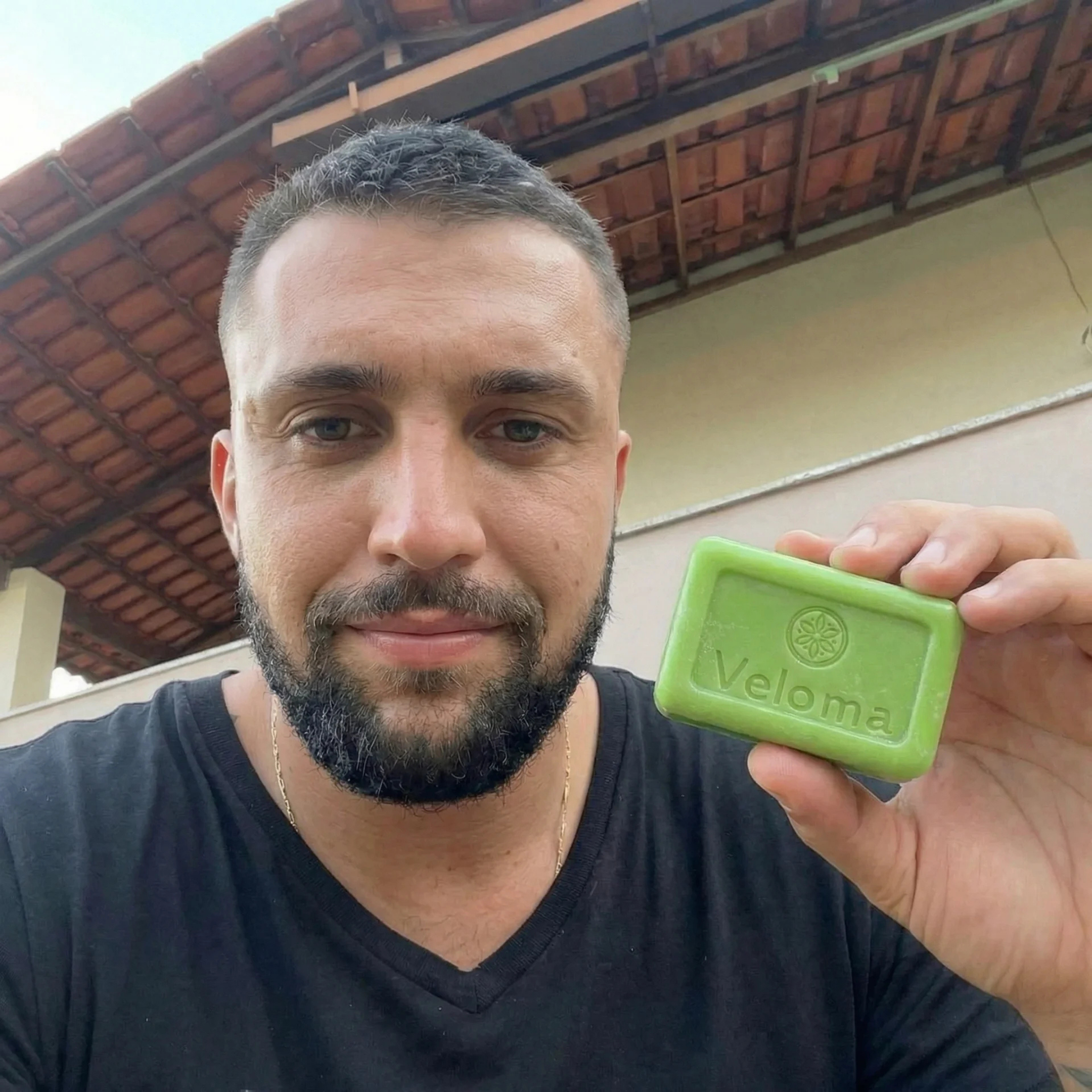 A man with a beard, wearing a black t-shirt, holds up a green bar of Veloma soap.