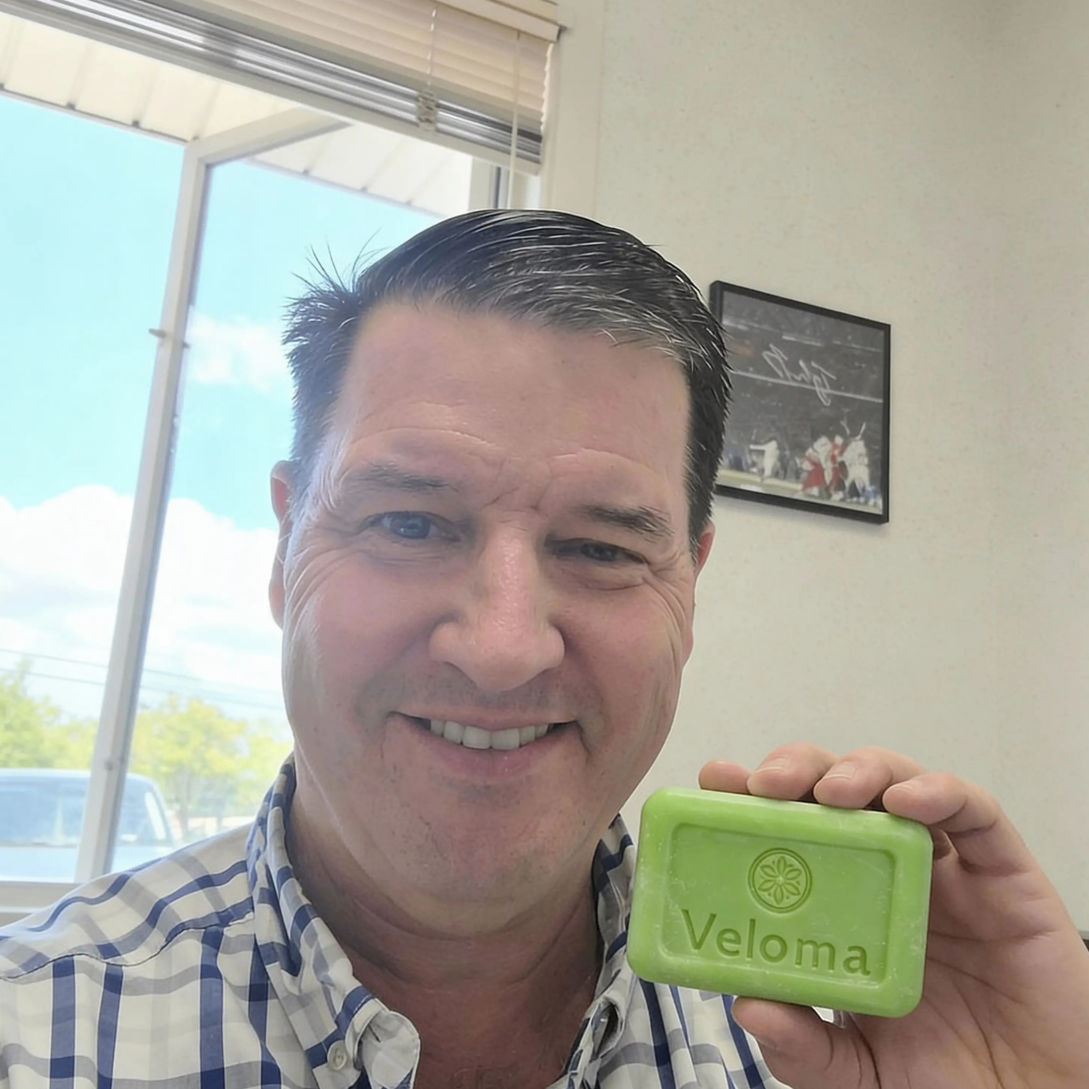 A smiling man in a plaid shirt holds up a green bar of Veloma soap.