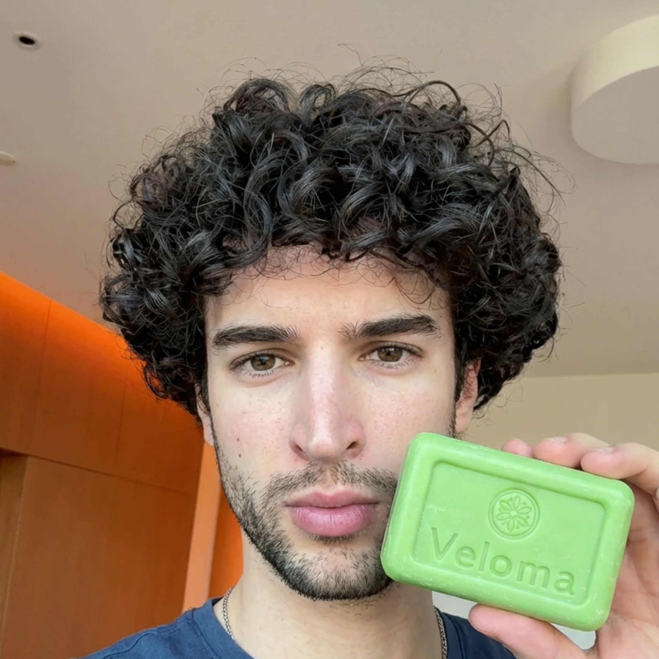 A young man with dark curly hair holds up a green bar of Veloma soap.