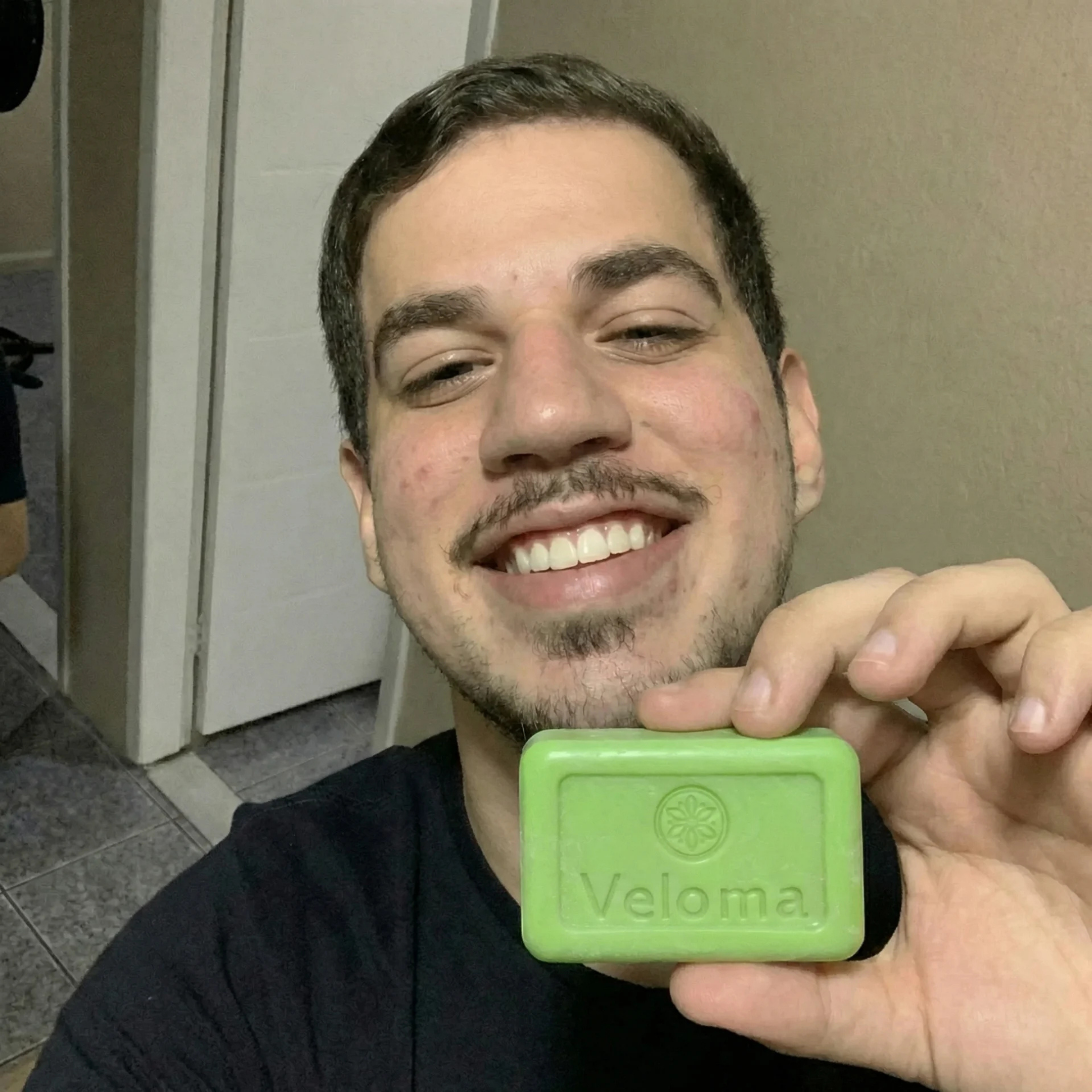 A smiling man holds up a green bar of soap with the brand name Veloma on it.