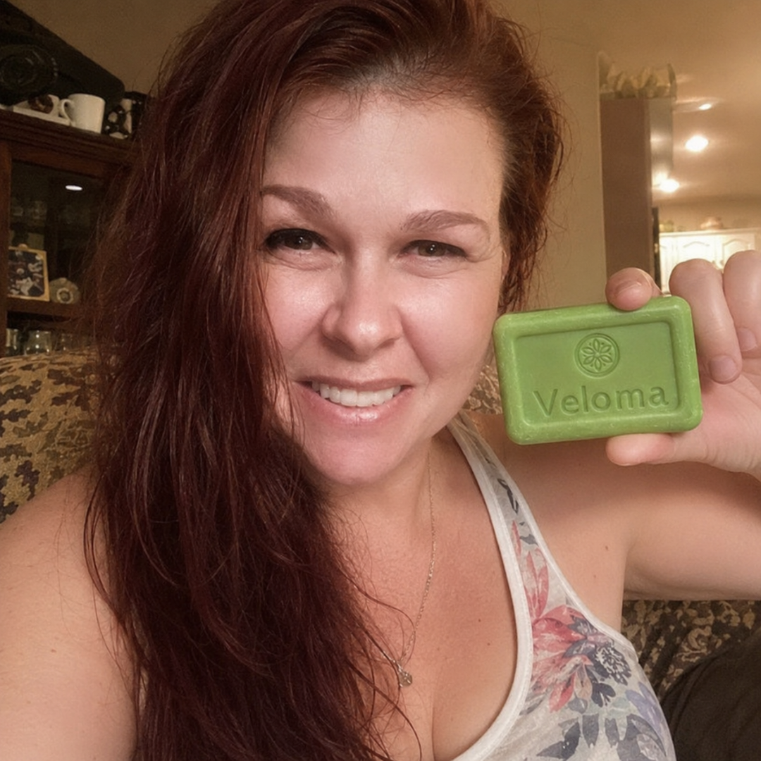 A smiling woman with long red hair holds up a green bar of Veloma soap.