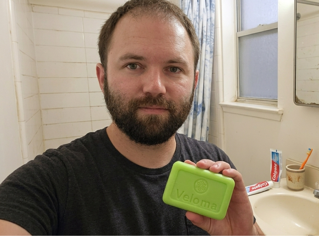 Bearded man in a grey t-shirt stands in a bathroom holding a bar of soap