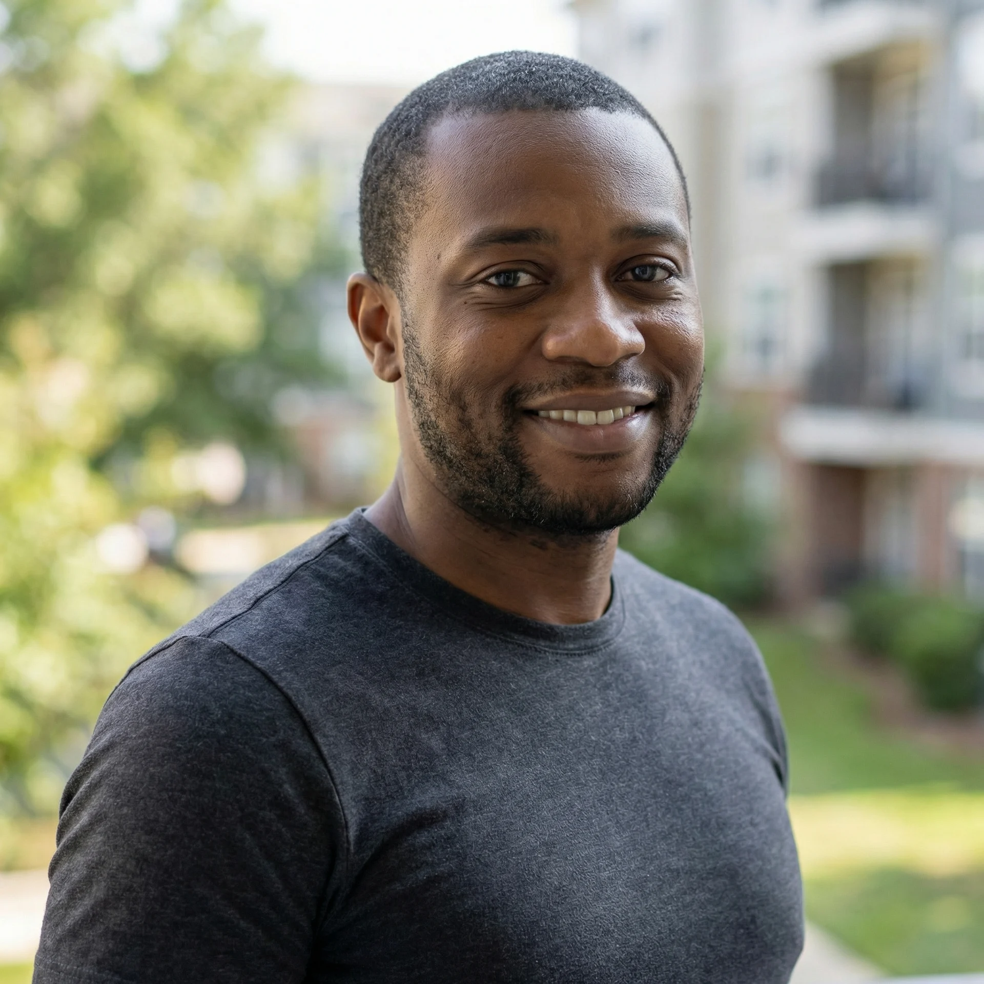 A Black man in a dark grey t-shirt smiles for a headshot outdoors.