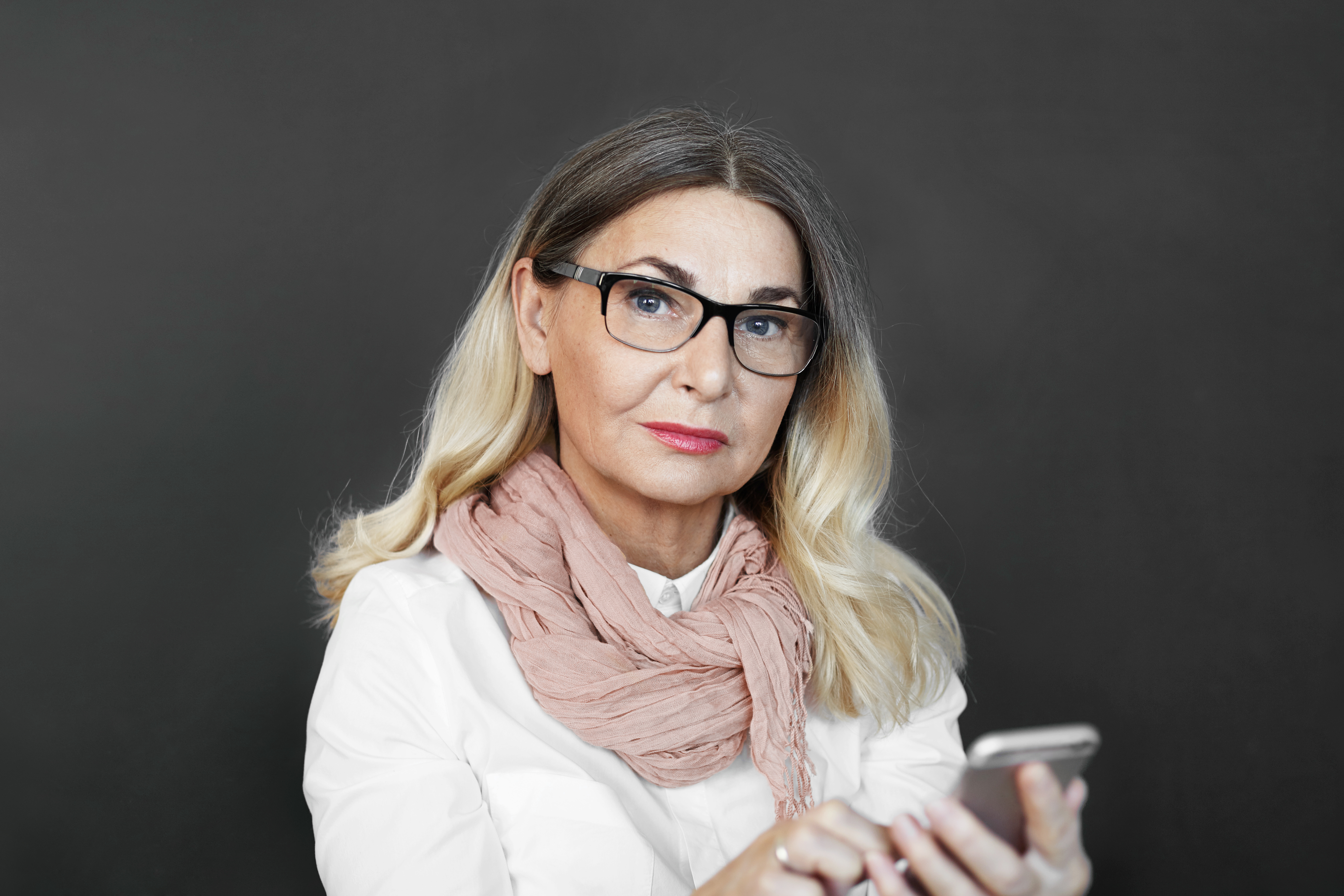 A woman with glasses and a pink scarf holds a smartphone while looking directly at the camera.