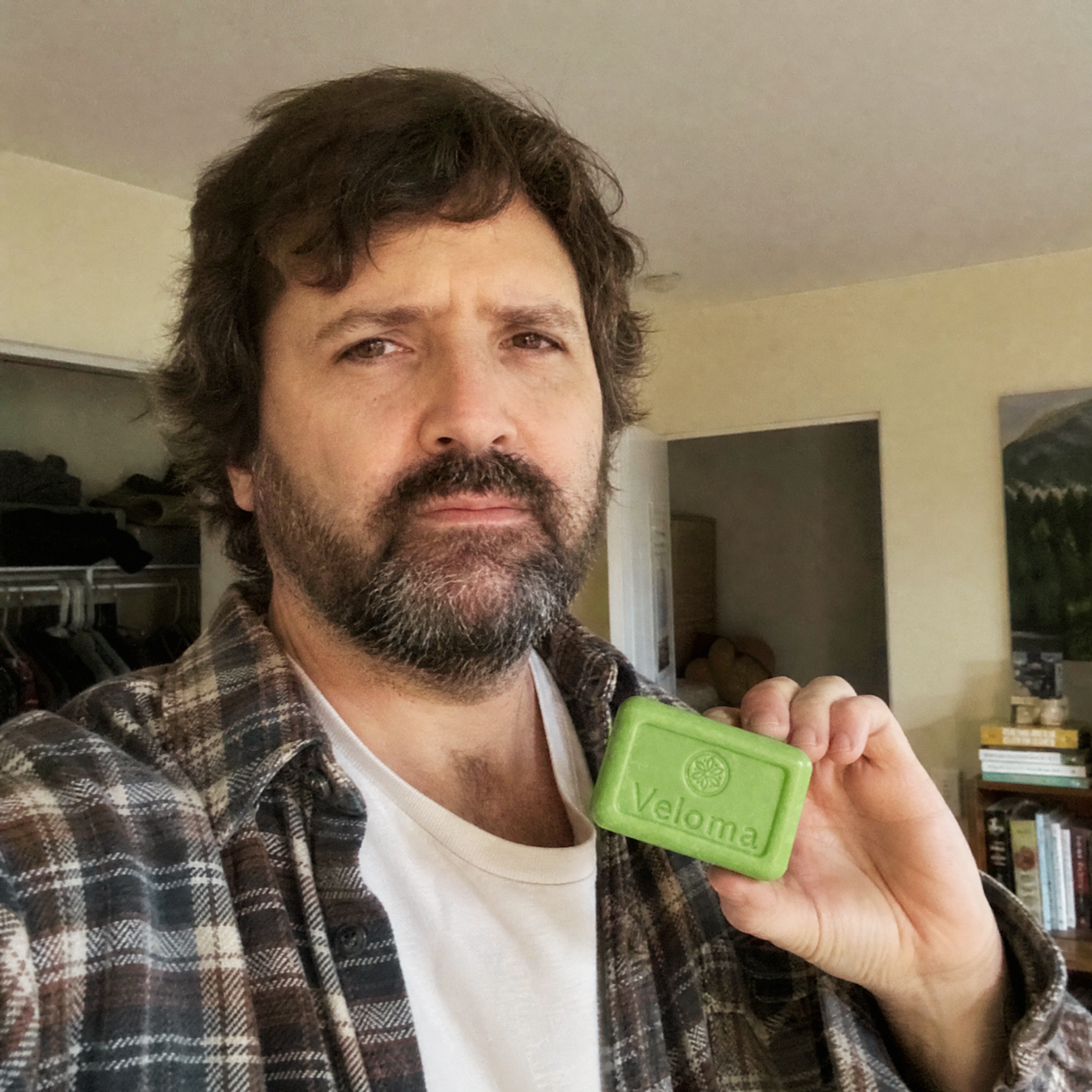 A man with a beard wearing a plaid shirt holds up a green bar of Veloma soap.