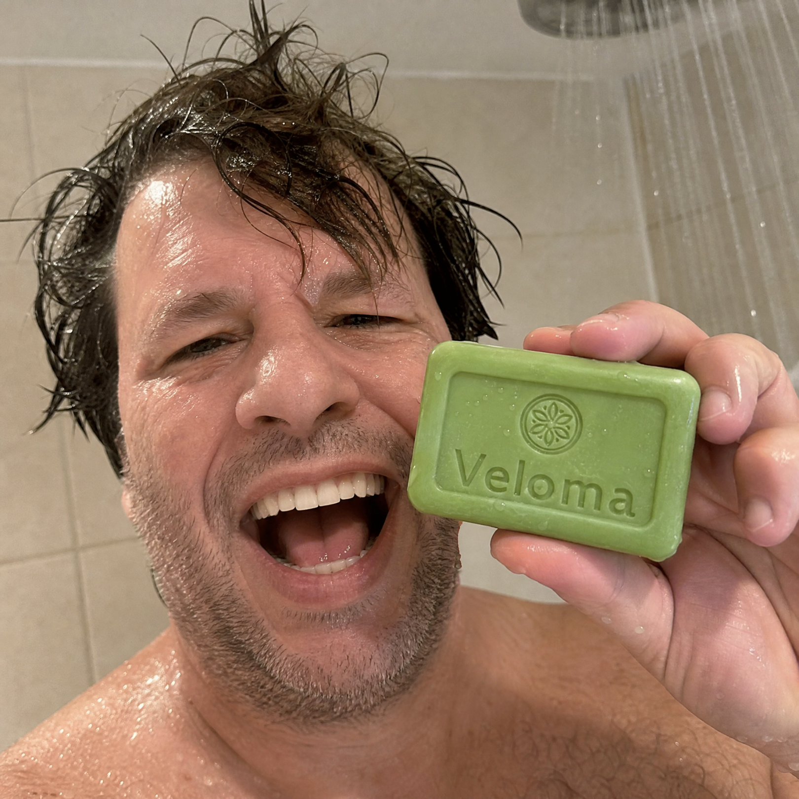 A happy man with wet hair in the shower holds up a green bar of Veloma soap.