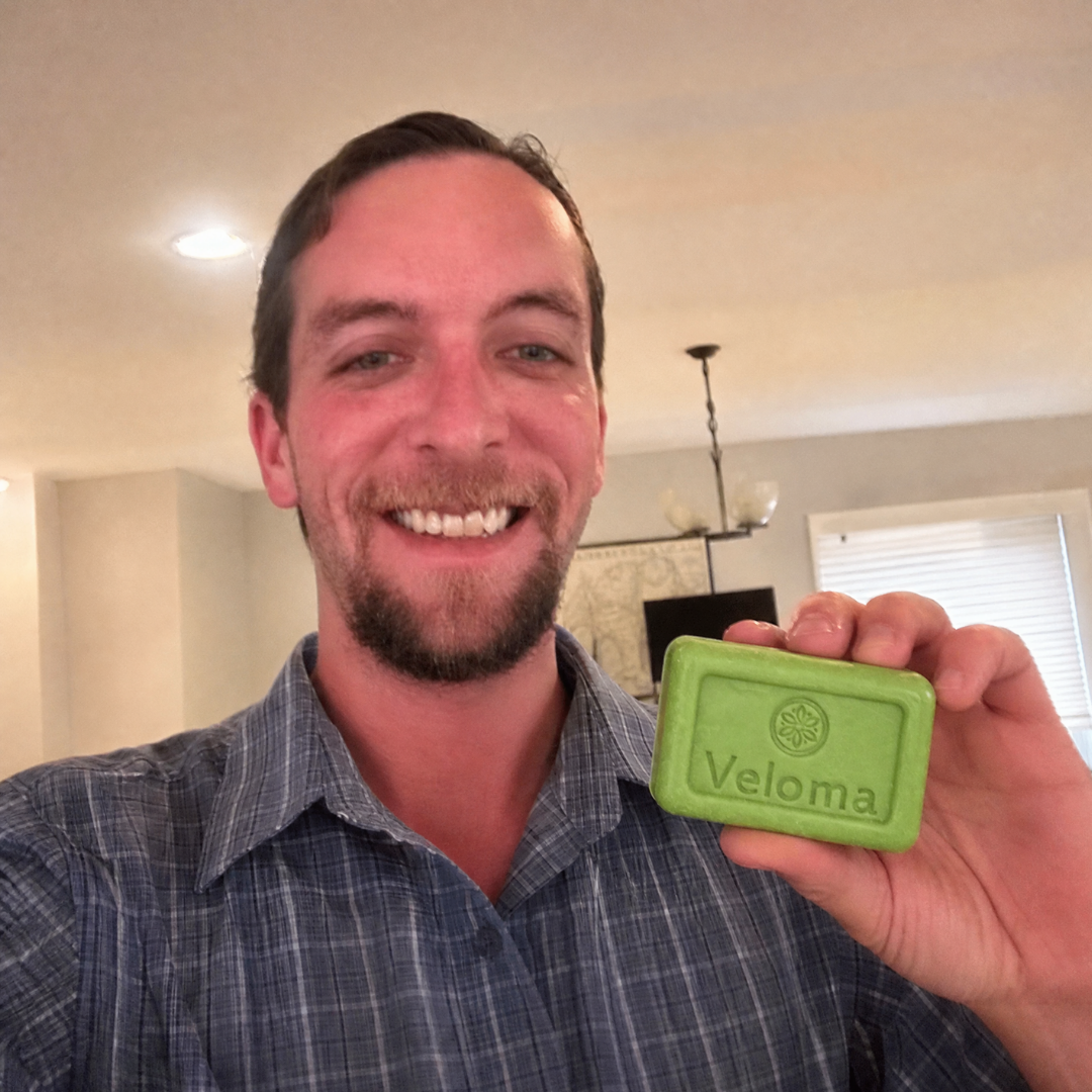A smiling man in a plaid shirt holds up a green bar of Veloma soap.