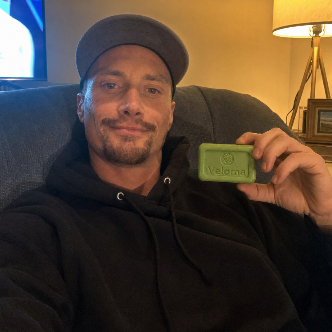 Man in a hoodie and cap holds up a bar of Veloma soap