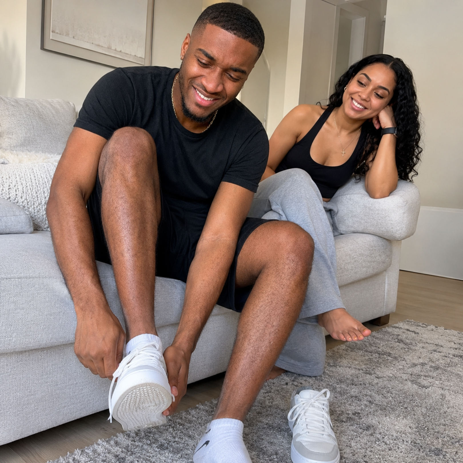 A smiling man sits on a couch putting on his shoes as a woman watches him.