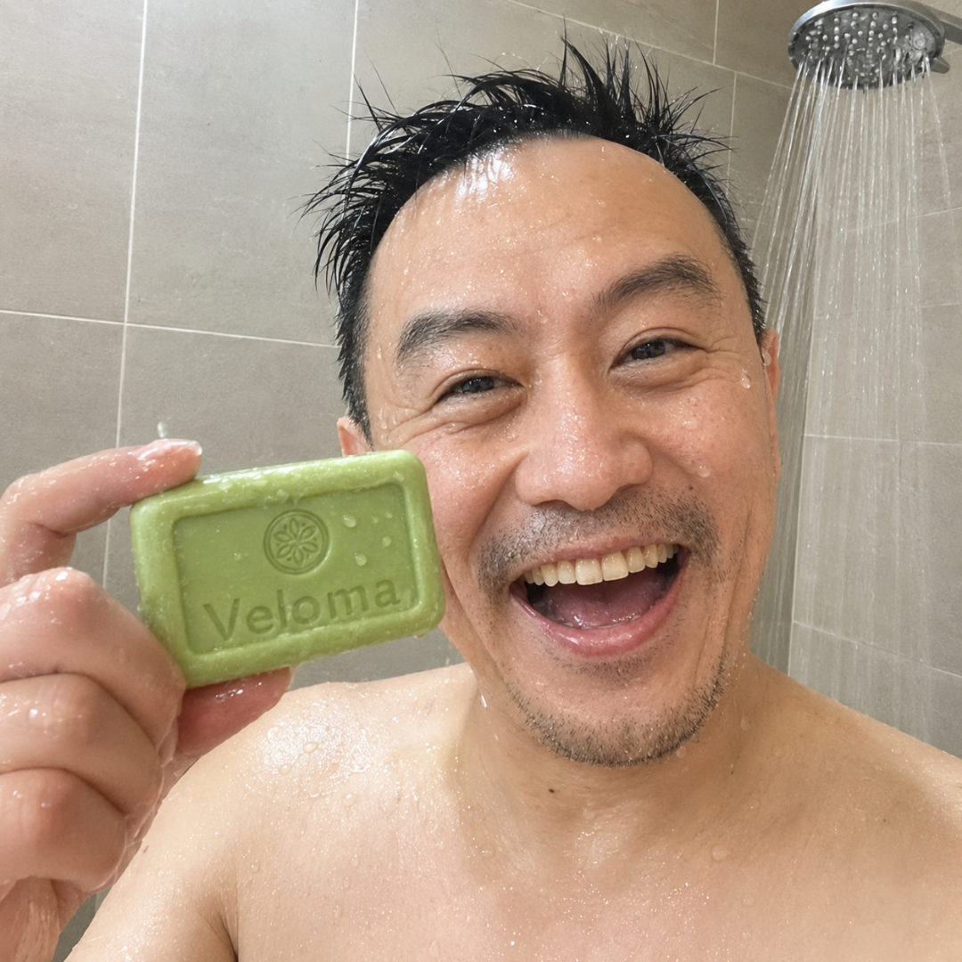 A smiling man in a shower holds up a green bar of Veloma soap to the camera.