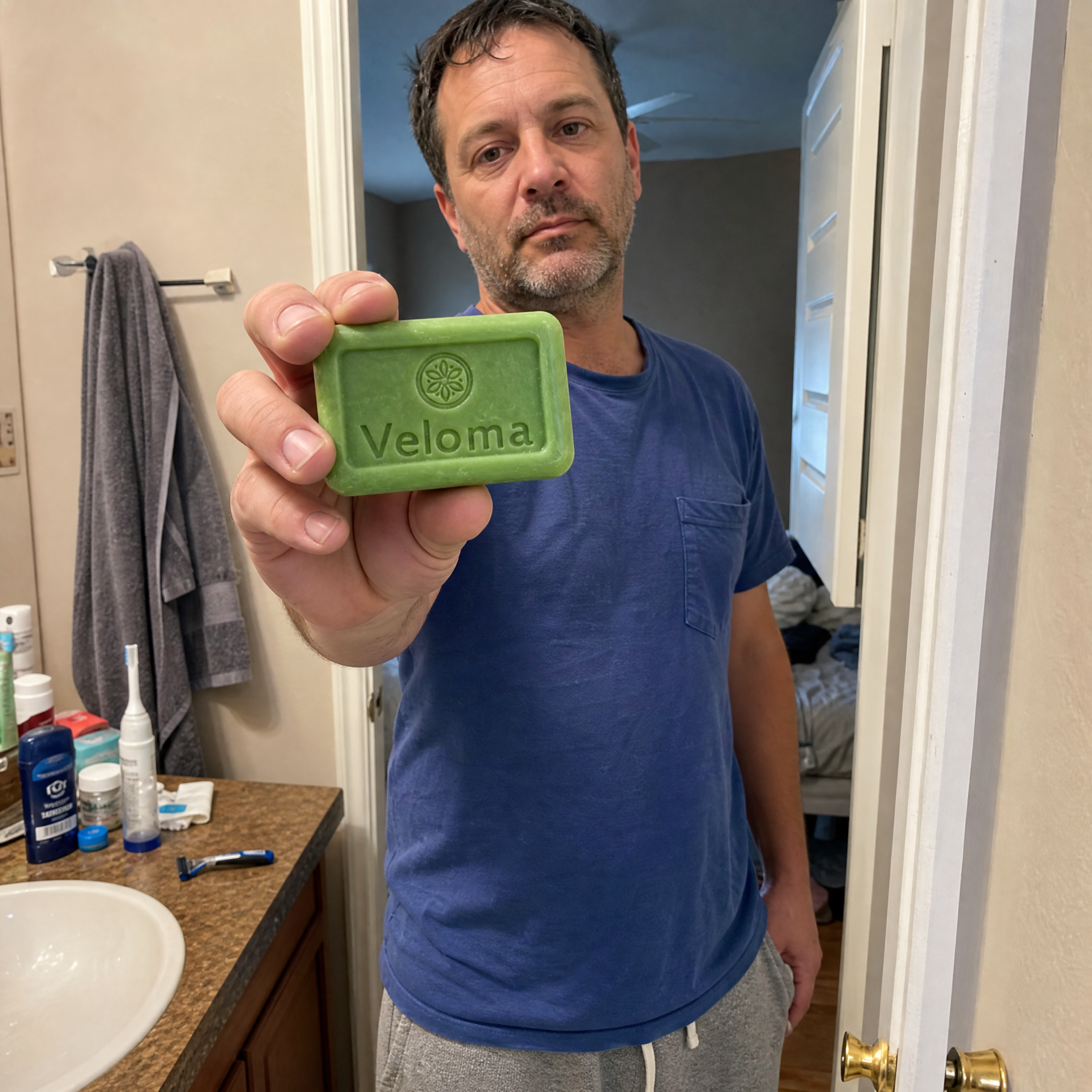 A man in a bathroom holds up a green bar of soap with the word Veloma on it.