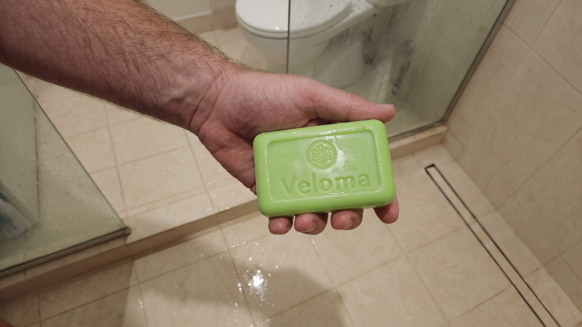 A man's hand holds a green bar of Veloma soap in a tiled shower.