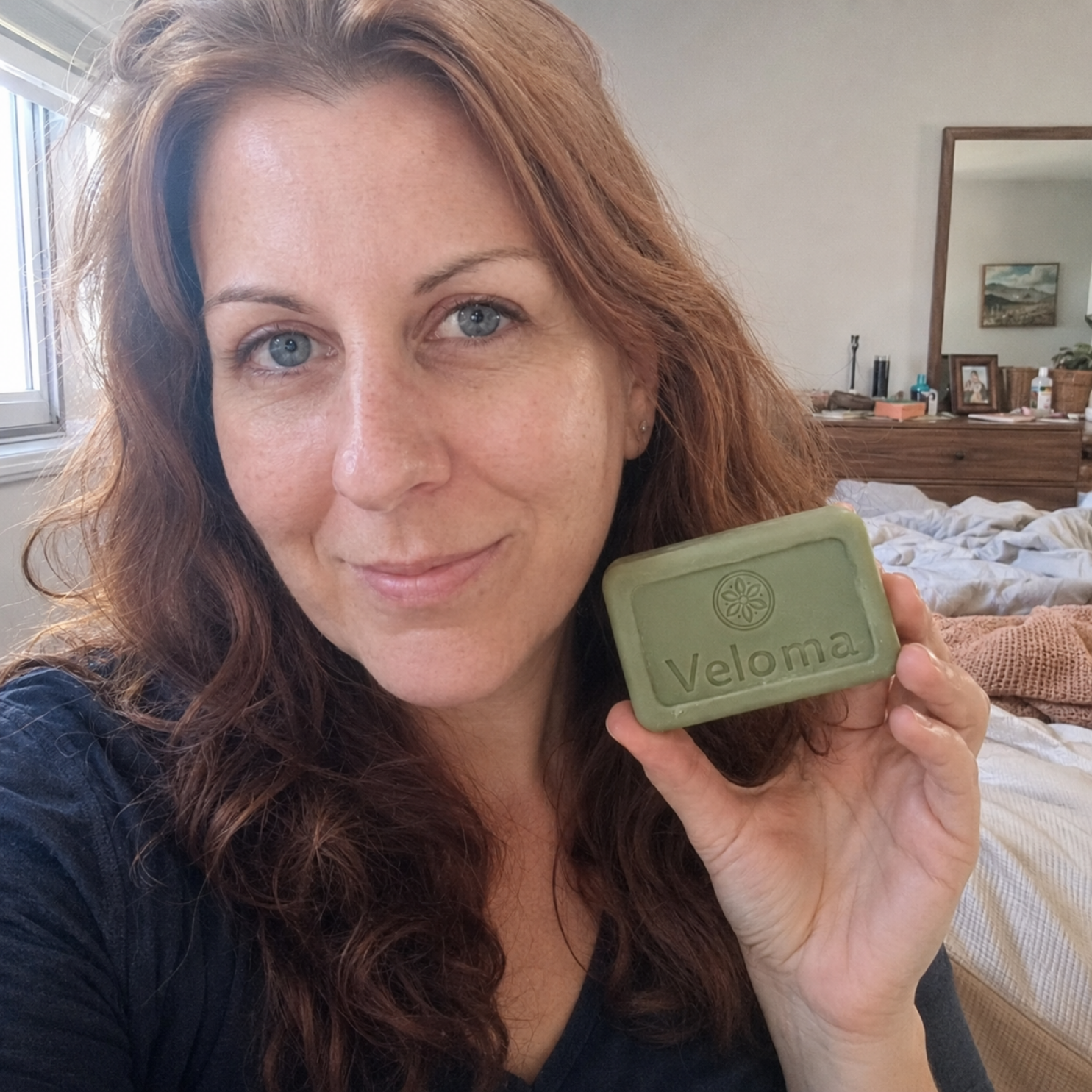 A woman with reddish-brown hair holds a green bar of soap labeled 'Veloma' in her bedroom.