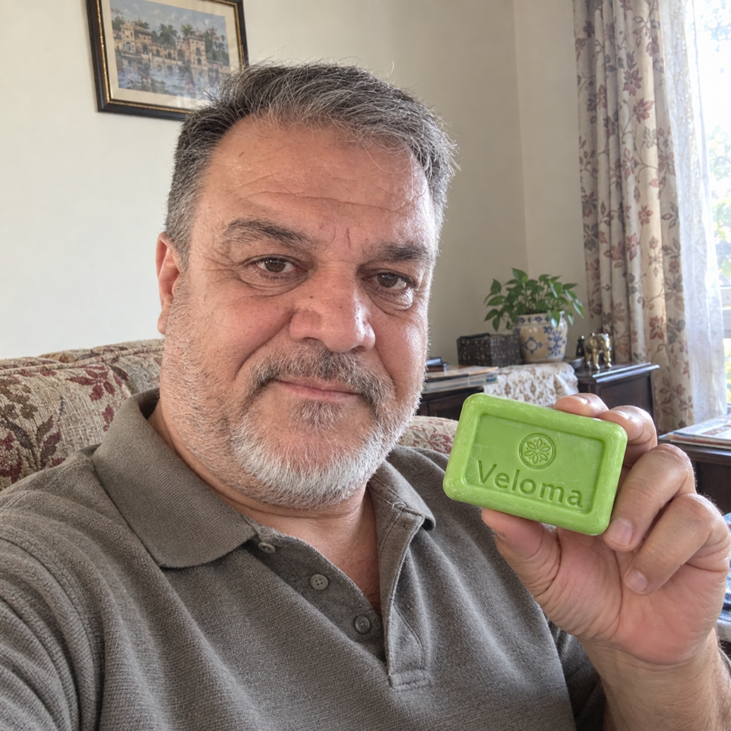 A middle-aged man in a gray polo shirt holds up a green bar of soap branded 'Veloma'.