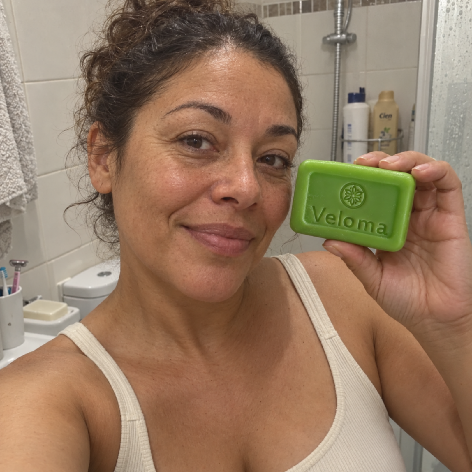 A woman with her hair up smiles while holding a green bar of Veloma soap in a bathroom.