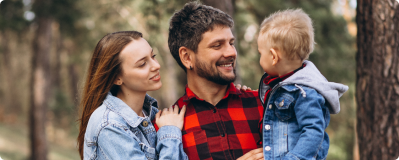 A young family of three, a man, woman, and toddler, smiling at each other in the woods.