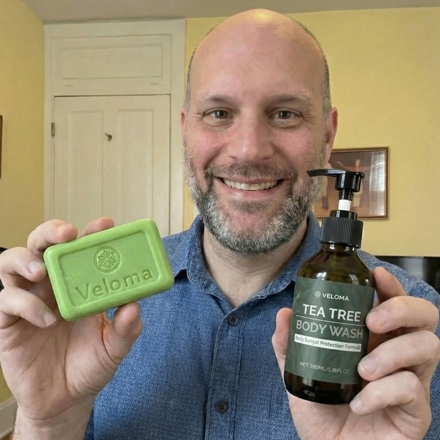 A smiling man holds a bar of Veloma soap and a bottle of Veloma Tea Tree Body Wash.