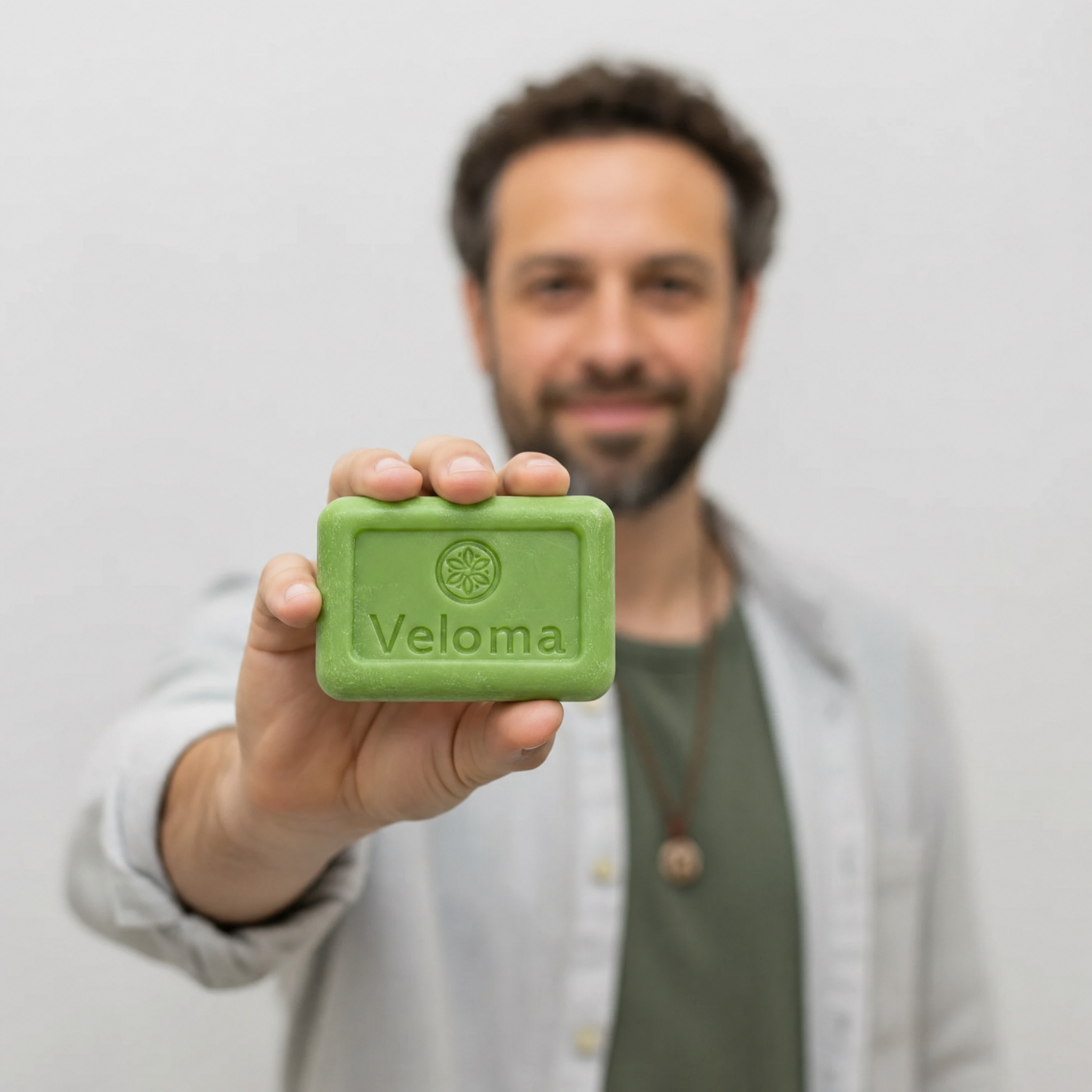 A man holds a green bar of Veloma soap towards the camera, with his face blurred behind.