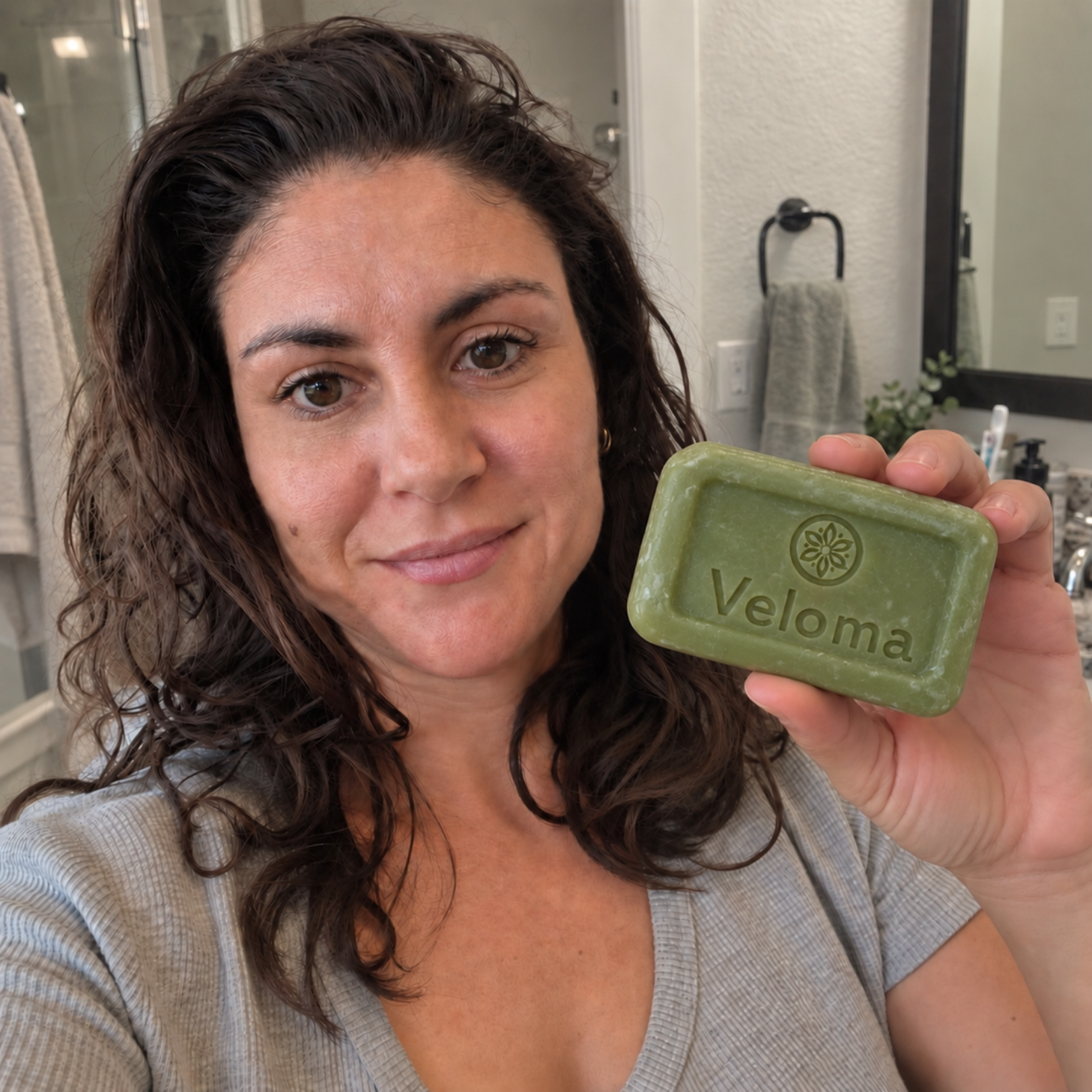 A smiling woman with dark hair holds up a green bar of soap with 'Veloma' written on it.