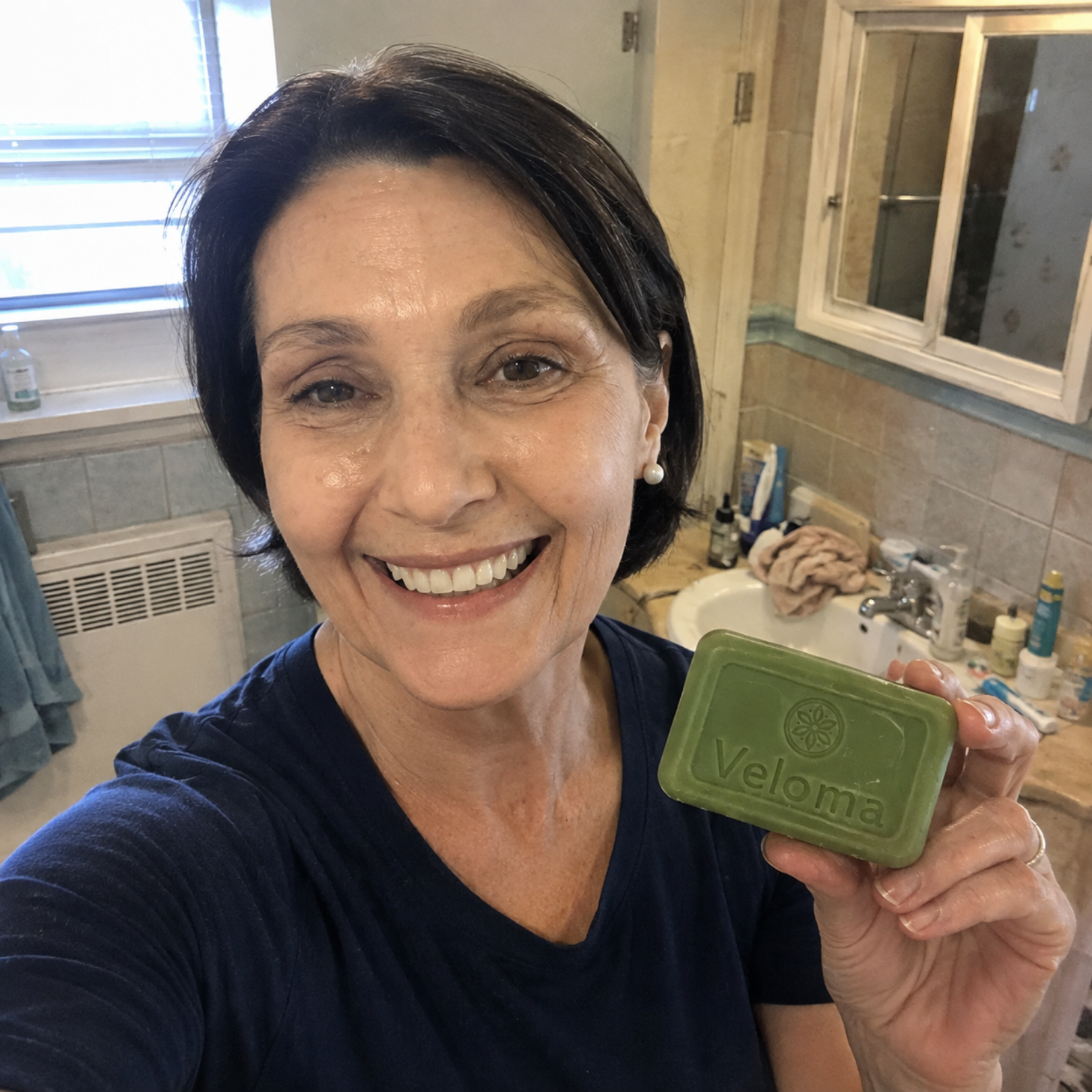 A smiling woman with dark hair holds up a green bar of Veloma soap in a bathroom.
