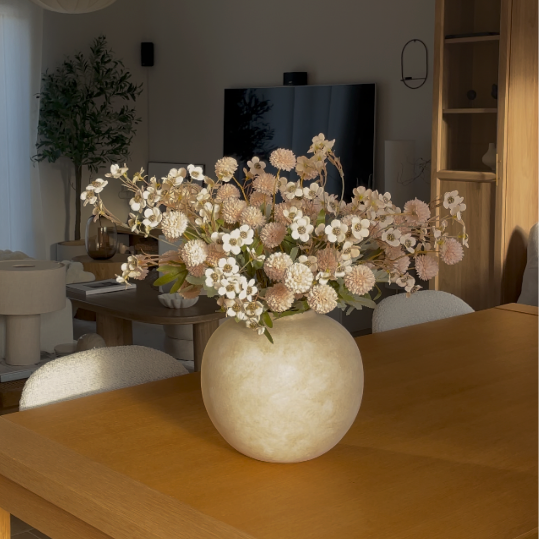 A round, cream-colored vase of pink and white flowers sits on a wooden table in a sunlit room.