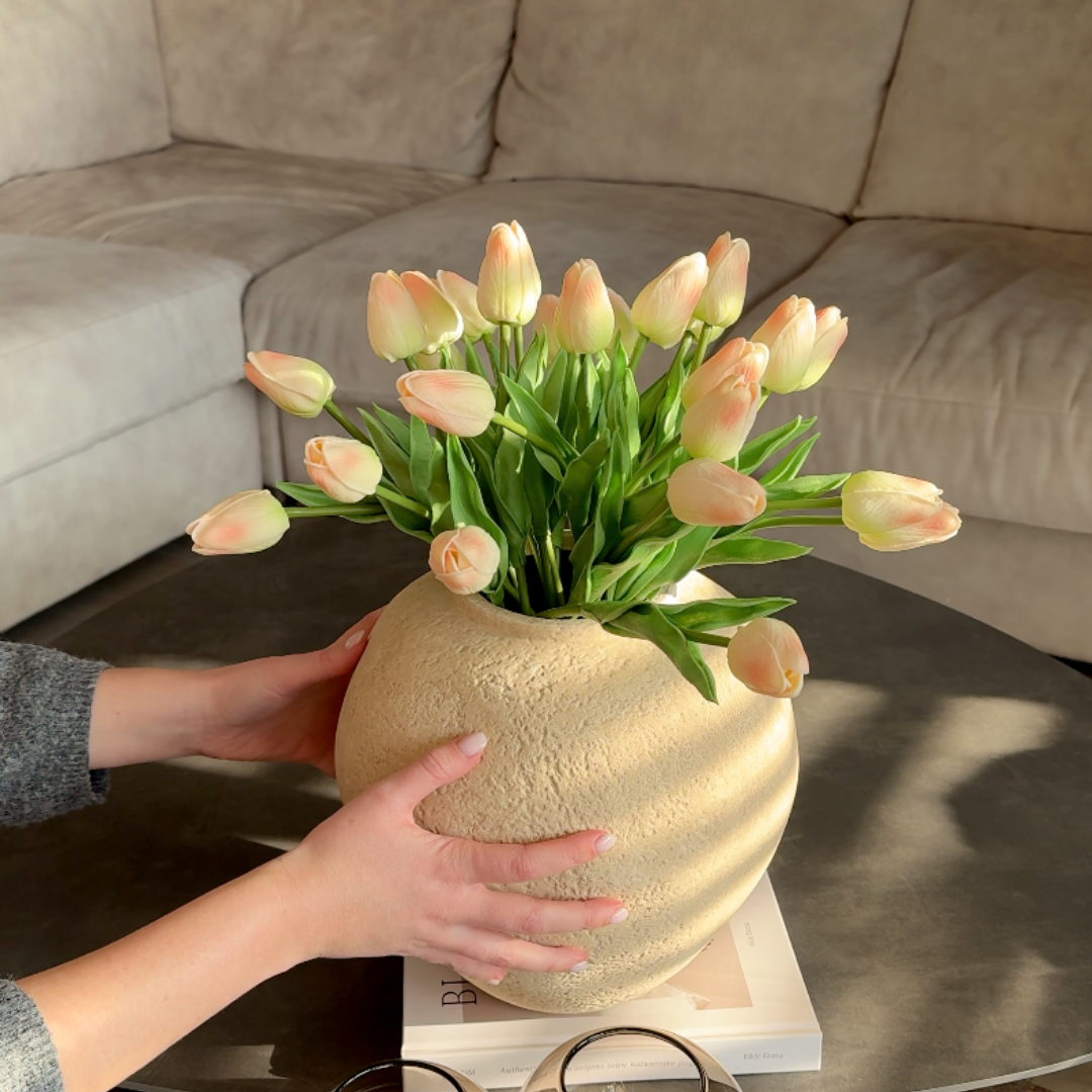 A person's hands hold a textured, light-colored vase filled with pink and white tulips on a table.