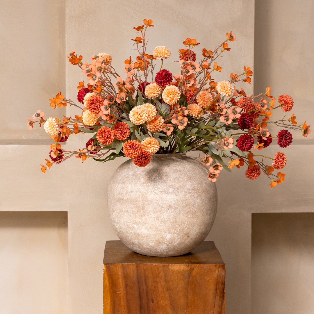 A bouquet of orange and red flowers in a large, round stone vase on a wooden pedestal.