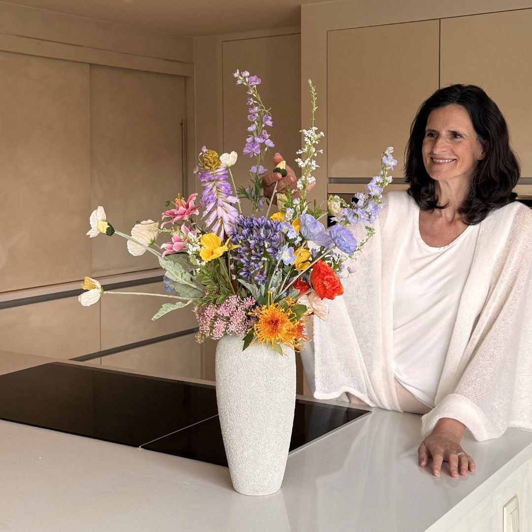 A woman in a white sweater smiles next to a large vase of colorful wildflowers on a kitchen island.