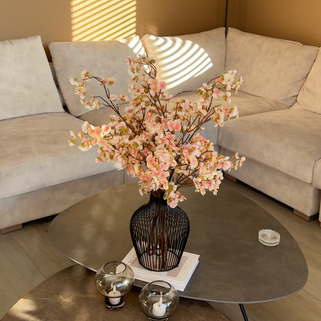 A black wire vase filled with pink blossoms on a coffee table in front of a sofa.