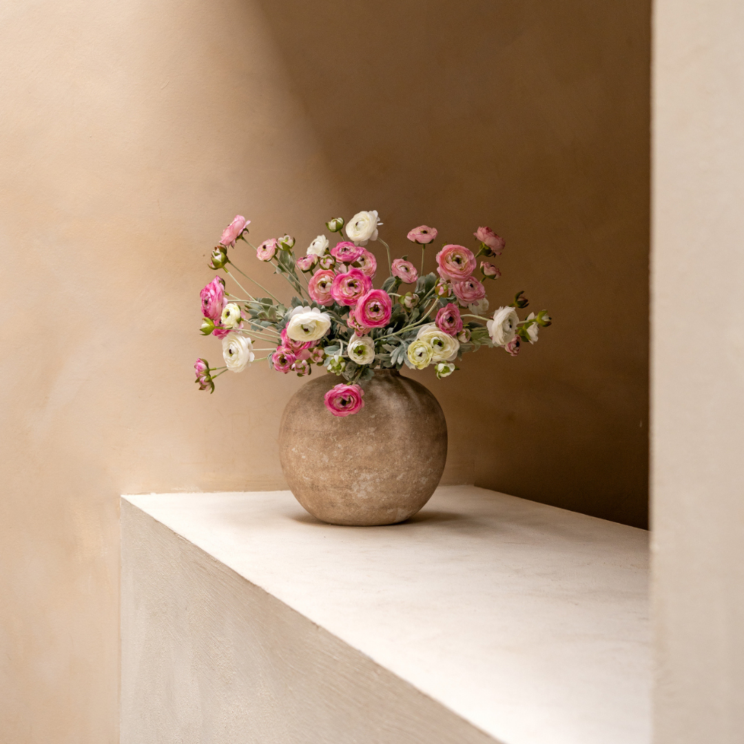 A round stone vase with pink and white ranunculus flowers sits on a ledge against a tan wall.