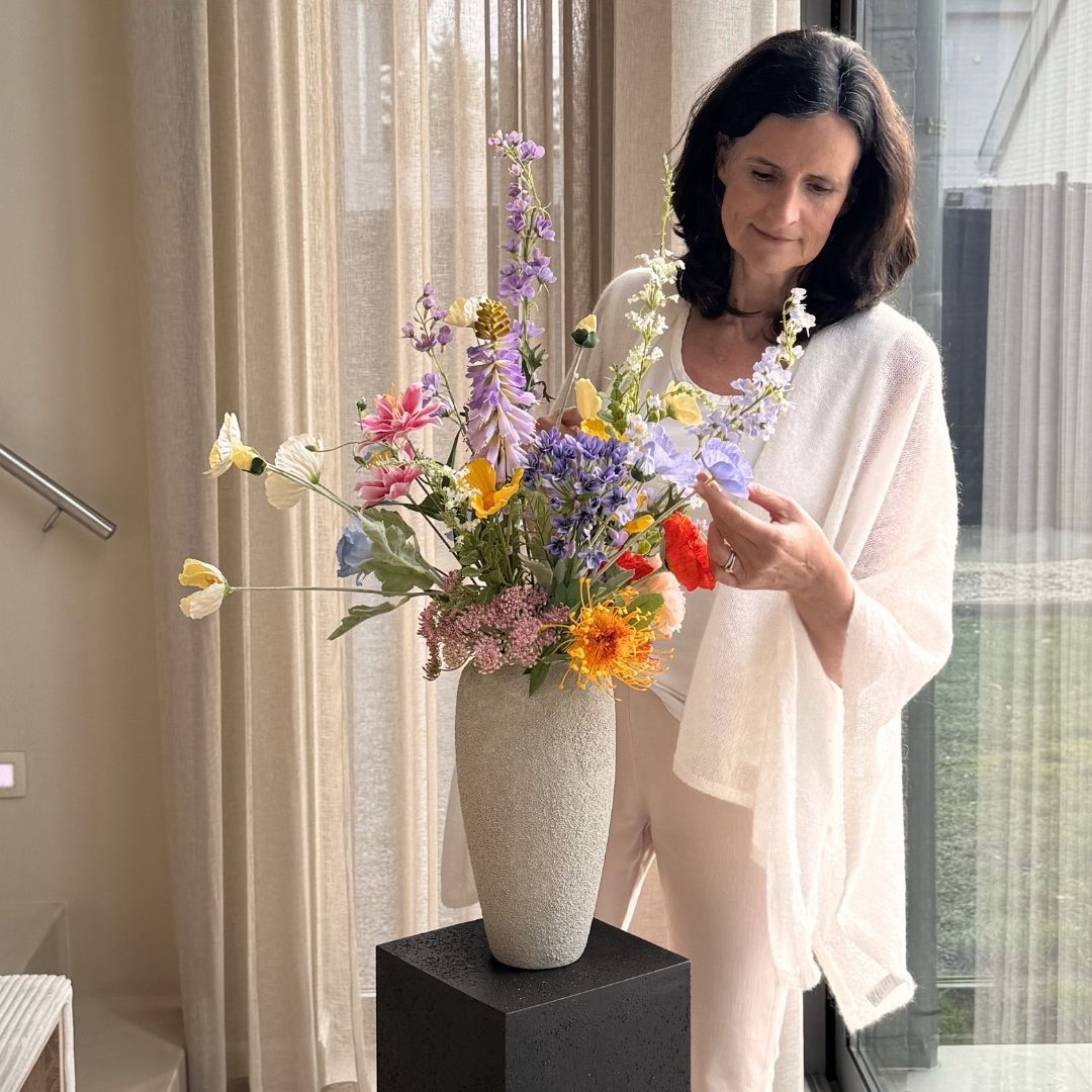 A woman in white clothing arranges a large, colorful bouquet of flowers in a tall grey vase.