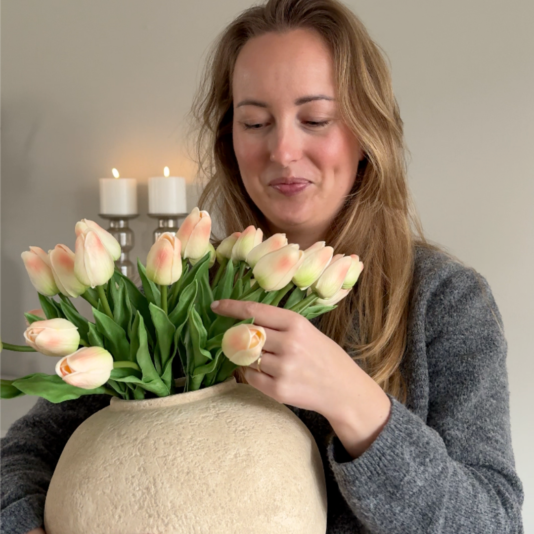 A woman in a grey sweater holds a large vase of light pink and cream tulips.