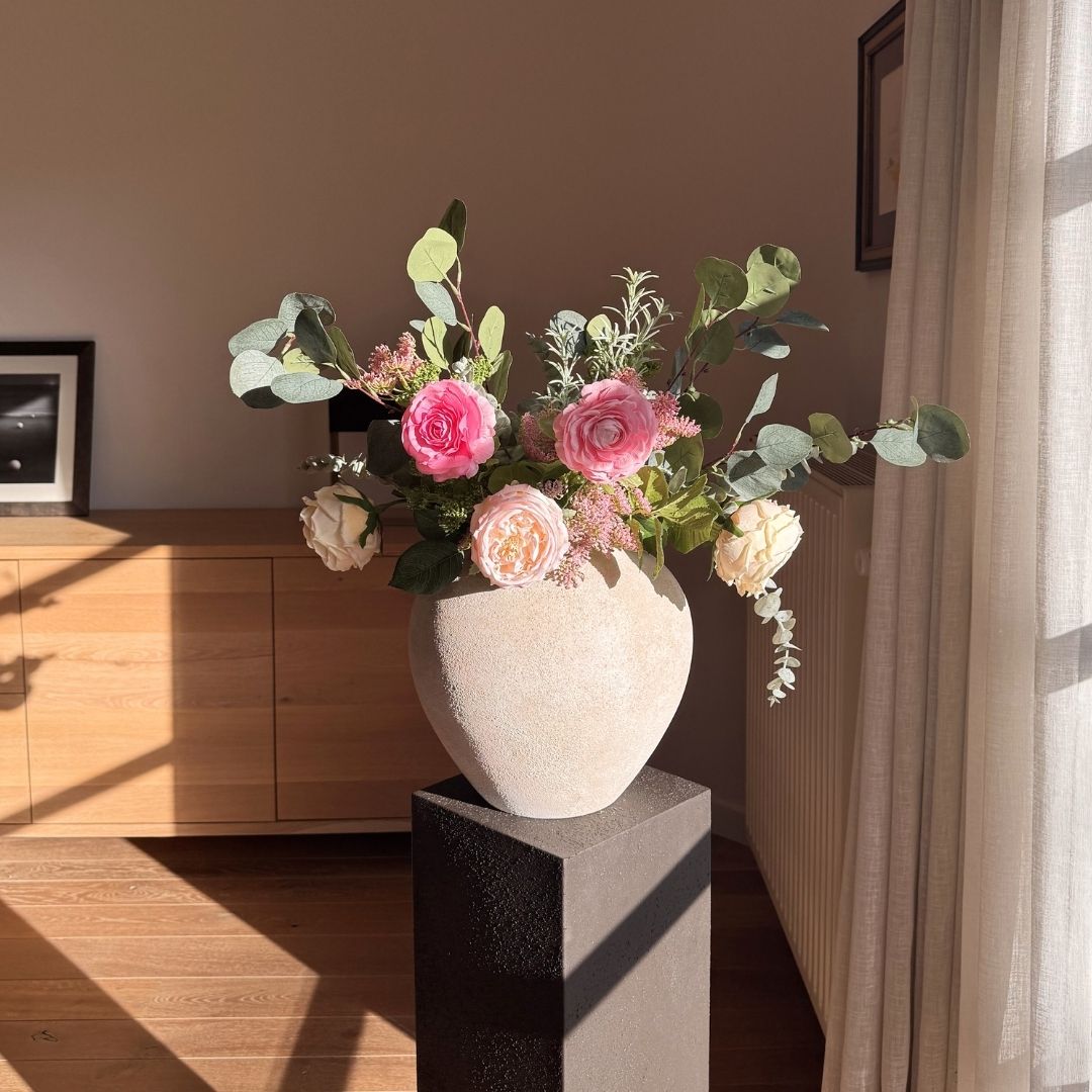 A textured vase of pink and cream roses and eucalyptus sits on a dark pedestal in a sunlit room.
