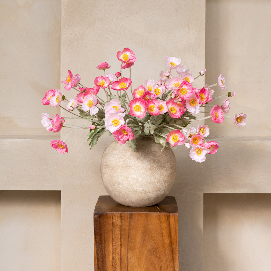 A bouquet of pink and white flowers with yellow centers in a round stone vase on a wooden stand.