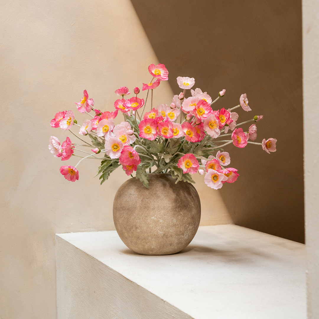 A bouquet of pink poppies with yellow centers in a spherical stone vase on a white ledge.
